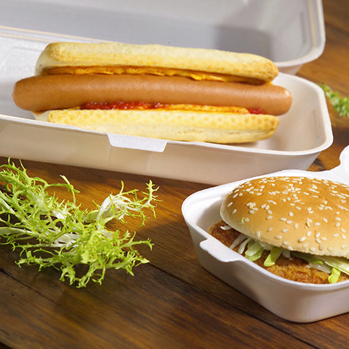Ein Hotdog mit Ketchup und Senf wird in einer Starpak 100 Allzweckbox mit Klappdeckel XPS 1800 ml serviert, daneben ein Sesam-Hamburger mit Salat in einer weiteren Starpak-Box. Auf dem Holztisch liegt etwas krauses Salatblatt.