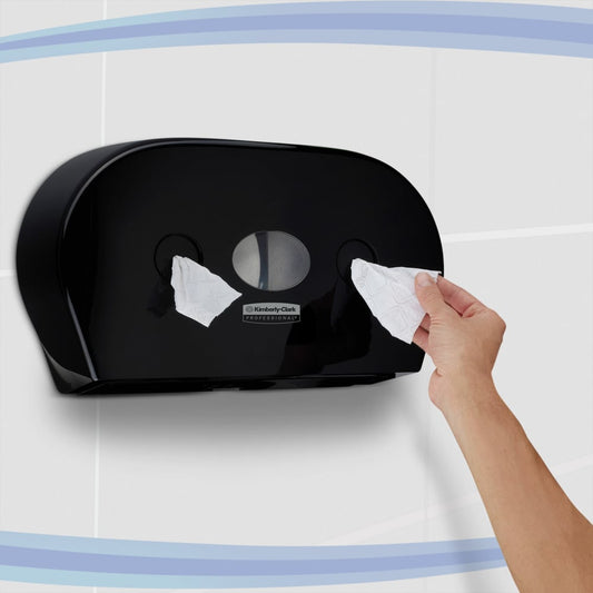 Eine Person zieht Toilettenpapier aus einem schwarzen Kimberly-Clark Professional™ Toilet Tissue Spender - Zentralentnahme (Jumbo) der Kimberly-Clark GmbH, der an einer gefliesten Wand mit blauen geschwungenen Streifen angebracht ist.