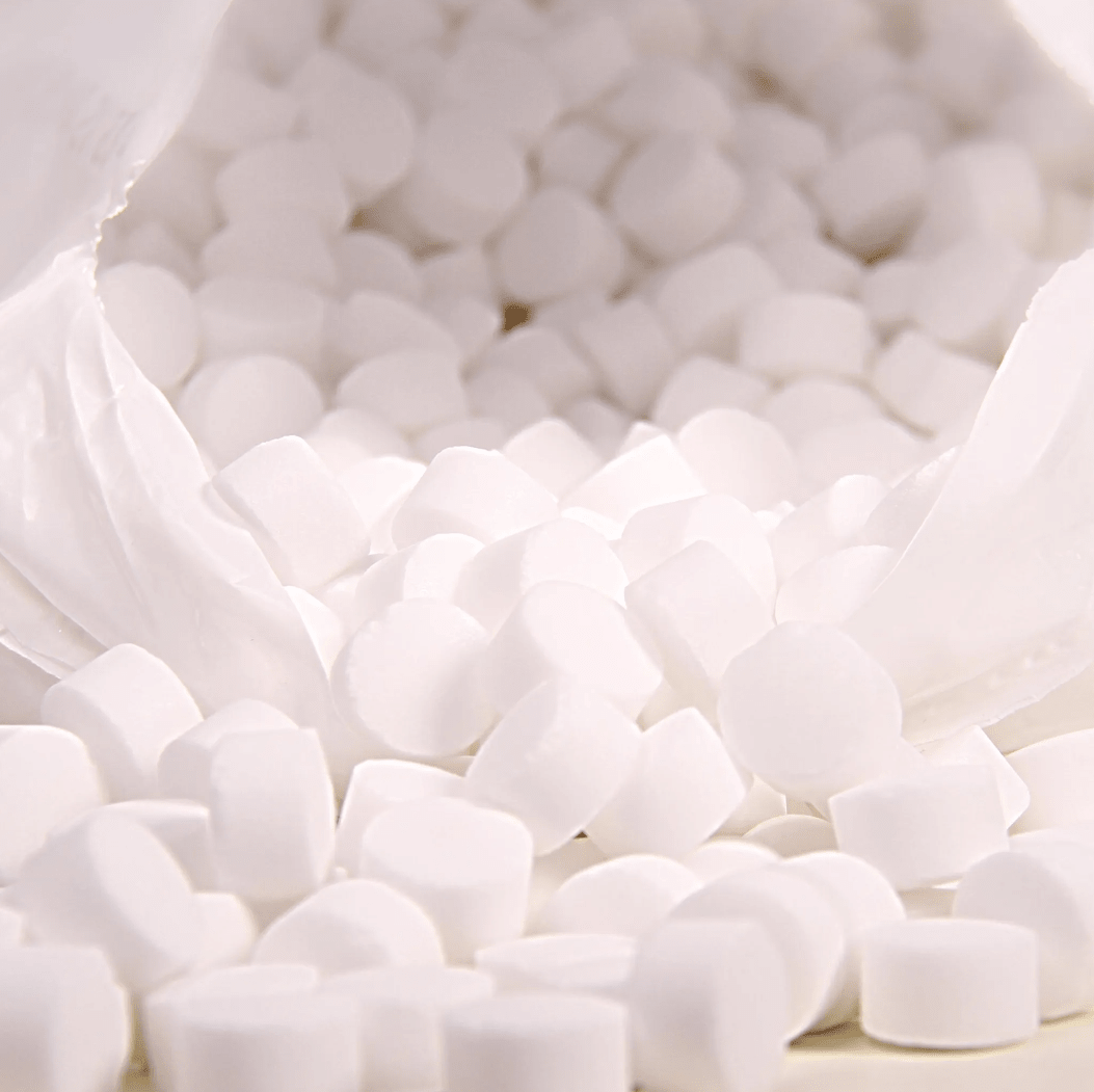 Na zbliżeniu widać białą plastikową torbę z napisem „K+S Minerals and Agriculture GmbH”, zawierającą tabletki soli warzonej Regenit zgodne z normą EN 973, typ | worek (25 kg), które spadają na powierzchnię. Te małe, okrągłe tabletki mają jednolity kształt i rozmiar oraz idealnie nadają się do stosowania w instalacjach zmiękczających wodę.