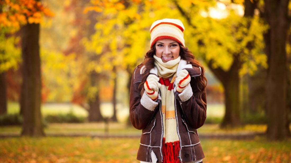 Frau in warmer Herbstkleidung mit Schal und Mütze lächelt im Park, umgeben von bunten Bäumen und herbstlicher Atmosphäre