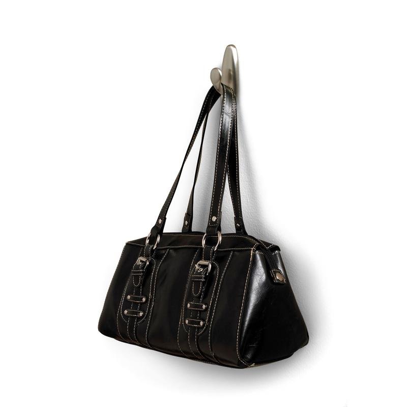 Na metalowym haku Command™ "klasycznym" firmy 3M Deutschland GmbH wisi czarna skórzana torebka z podwójnymi uchwytami i ozdobnymi klamrami. Torebka ma białe szwy i lekko błyszczącą powierzchnię.