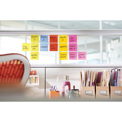 Rząd karteczek 3M Post-it®, żółte, 76x76 mm (90 arkuszy/blok, 6 bloków/opakowanie) jest ułożony na szklanym dzielniku; poniżej na biurku znajdują się segregatory, książki, butelka wody i inne przybory.