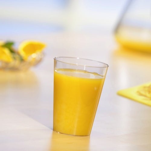 Kubek Starpak 10, PS 0,2l przezroczysty, wypełniony sokiem pomarańczowym stoi na jasnym stole. W tle widać niewyraźne plasterki pomarańczy i dzbanek.