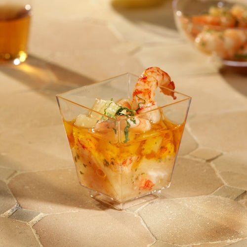 Miseczka Starpak 50 Fingerfood (PS glasklar) od Starpak, wypełniona krewetkami i koktajlem z owoców morza, udekorowana ziołami, stoi na beżowej, teksturowanej powierzchni. Inne kubki są rozmyte w tle – idealne na stylowe wydarzenia.