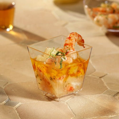 Miseczka Starpak 50 Fingerfood (PS glasklar) od Starpak, wypełniona krewetkami i koktajlem z owoców morza, udekorowana ziołami, stoi na beżowej, teksturowanej powierzchni. Inne kubki są rozmyte w tle – idealne na stylowe wydarzenia.
