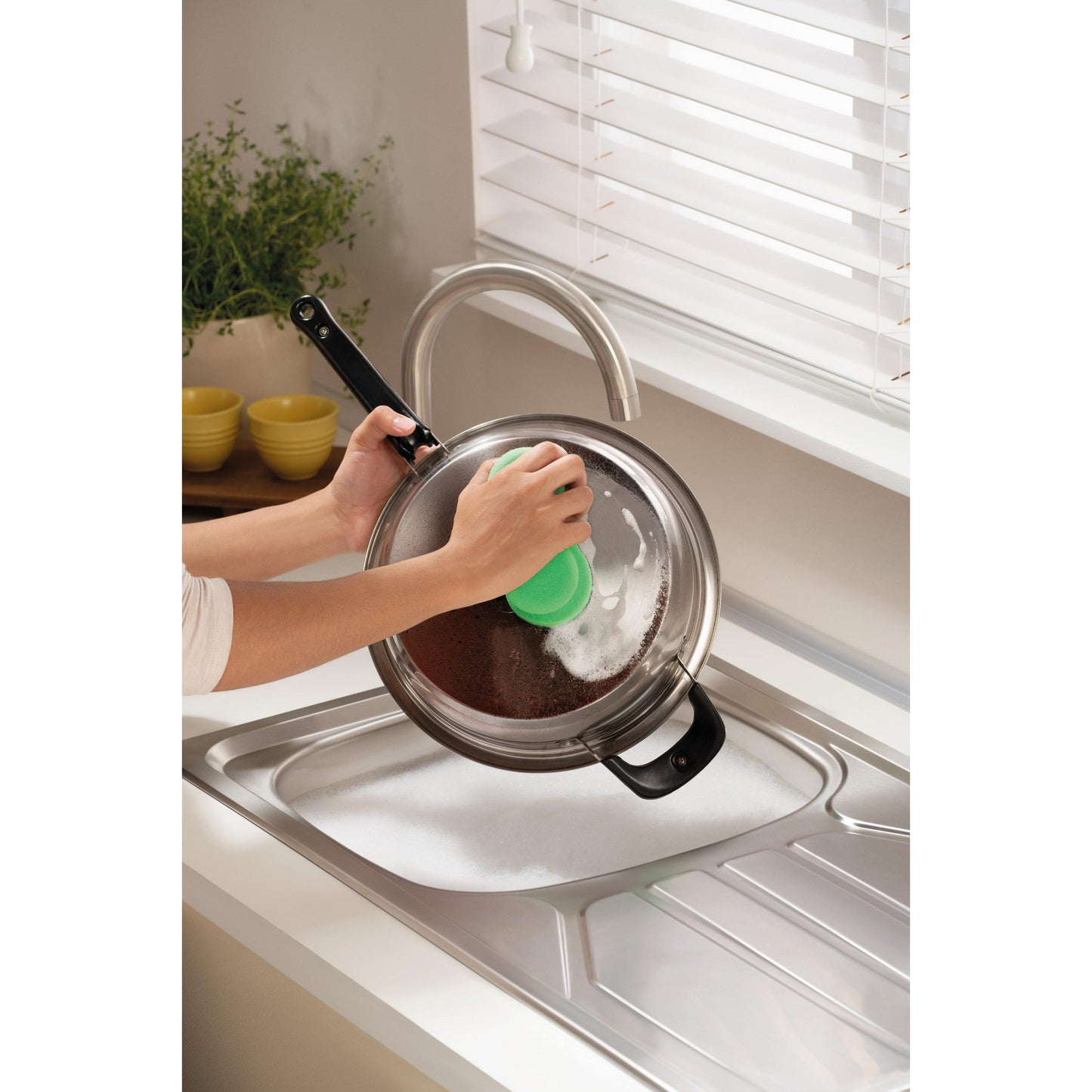 Ktoś czyści metalową pokrywkę garnka nad mydlaną umywalką za pomocą Scotch-Brite® Classic gąbki do czyszczenia 3M Deutschland GmbH, w pobliżu okna z białymi żaluzjami i blatem z rośliną i ułożonymi żółtymi miskami.