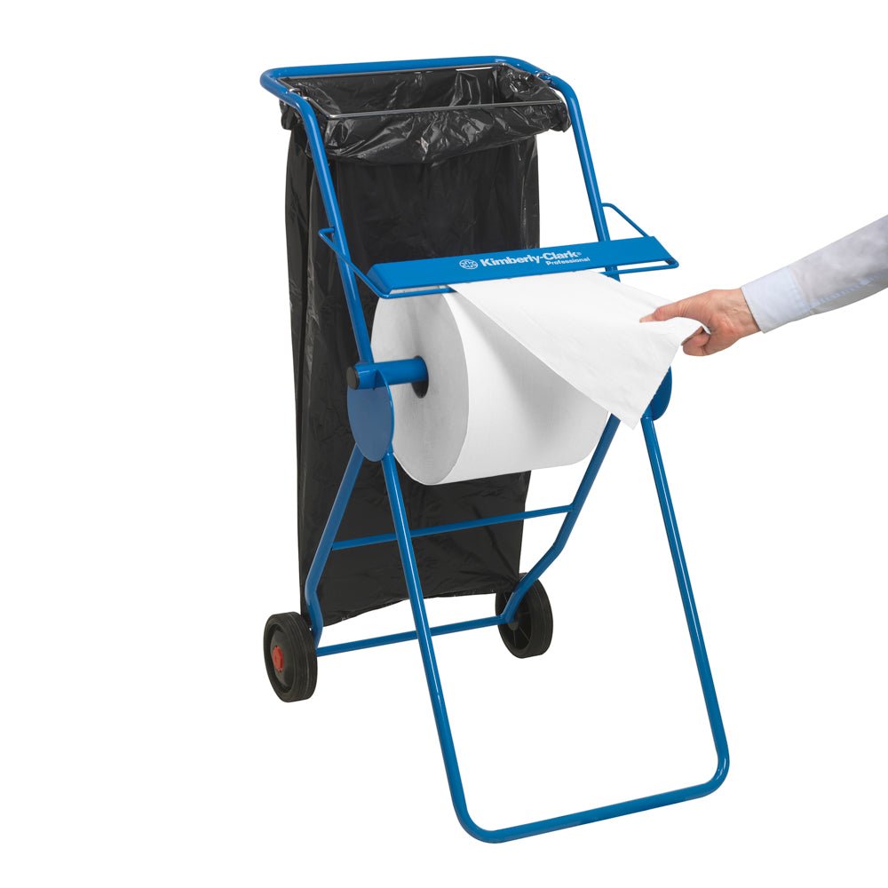 Osoba wyciąga papierowy ręcznik WypAll® L30 z ekstra szerokiej rolki (1000 arkuszy, biała) na niebieskim stojaku z rolkami od Kimberly-Clark GmbH; z tyłu przymocowana jest czarna plastikowa torba do zadań czyszczenia przemysłowego.