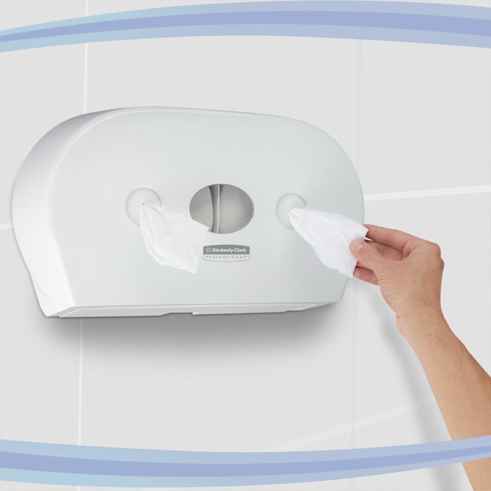 Ręka wyciąga chusteczki z dozownika papieru toaletowego Aquarius™ - centralny / biały / Jumbo firmy Kimberly-Clark GmbH, który jest wyposażony w dwie rolki i zamontowany na wyłożonej płytkami ścianie.