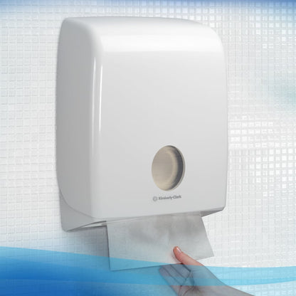 Ręka wyciąga ręcznik Scott® ESSENTIAL™ od Kimberly-Clark GmbH z białego, zamontowanego na ścianie dozownika w wyłożonej płytkami toalecie; 15 paczek po 340 arkuszy na karton.