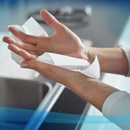 Osoba wyciera ręce ręcznikami Scott® ESSENTIAL™ Interfold (Białe, małe, 15 x 340 arkuszy) od Kimberly-Clark GmbH nad umywalką, w tle widać część blatu kuchennego i kranu.