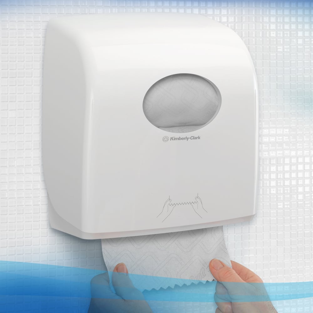Osoba wyciąga ręcznik z białego dozownika ręczników Scott® Essential™ firmy Kimberly-Clark GmbH z małym owalnym okienkiem, przez które można zobaczyć rolkę papieru wewnątrz, z teksturowanymi płytkami w tle. 6 białych rolek na karton.