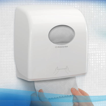 Osoba wyciąga ręcznik z białego dozownika ręczników Scott® Essential™ firmy Kimberly-Clark GmbH z małym owalnym okienkiem, przez które można zobaczyć rolkę papieru wewnątrz, z teksturowanymi płytkami w tle. 6 białych rolek na karton.
