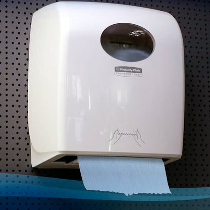 Biały, montowany na ścianie dozownik od Kimberly-Clark GmbH trzyma rolkę ręczników Scott® (304,00 m x 19,80 cm) z niebieskim ręcznikiem i prostą, zilustrowaną instrukcją na ciemnym tle z blachy perforowanej.