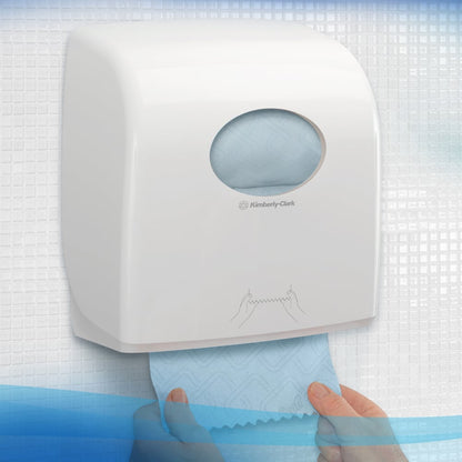 Osoba wyciąga niebieską rolkę ręczników Scott® Essential™ z białego dozownika Kimberly-Clark GmbH, zamontowanego na kafelkowanej ścianie z okrągłym oknem, które pokazuje znajdujące się w środku ręczniki oraz ilustrację wydawania ręczników.