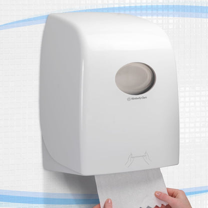 Dozownik ręczników w rolce Aquarius™ - Biały od Kimberly-Clark GmbH jest montowany na ścianie na płytkach, podczas gdy ręka wyciąga ręcznik od dołu. Jest idealny do miejsc publicznych i ma okrągłe okienko na górze oraz jasną instrukcję użytkowania poniżej.