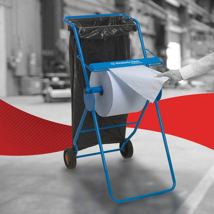 Osoba wyciąga ręcznik papierowy WypAll® L10 do powierzchni - duża rolka (38,00 x 23,50 cm) od Kimberly-Clark GmbH z niebieskiego metalowego stojaka z kółkami i przymocowanym do niego czarnym workiem na śmieci, co wskazuje na efektywne czyszczenie powierzchni w środowiskach przemysłowych.