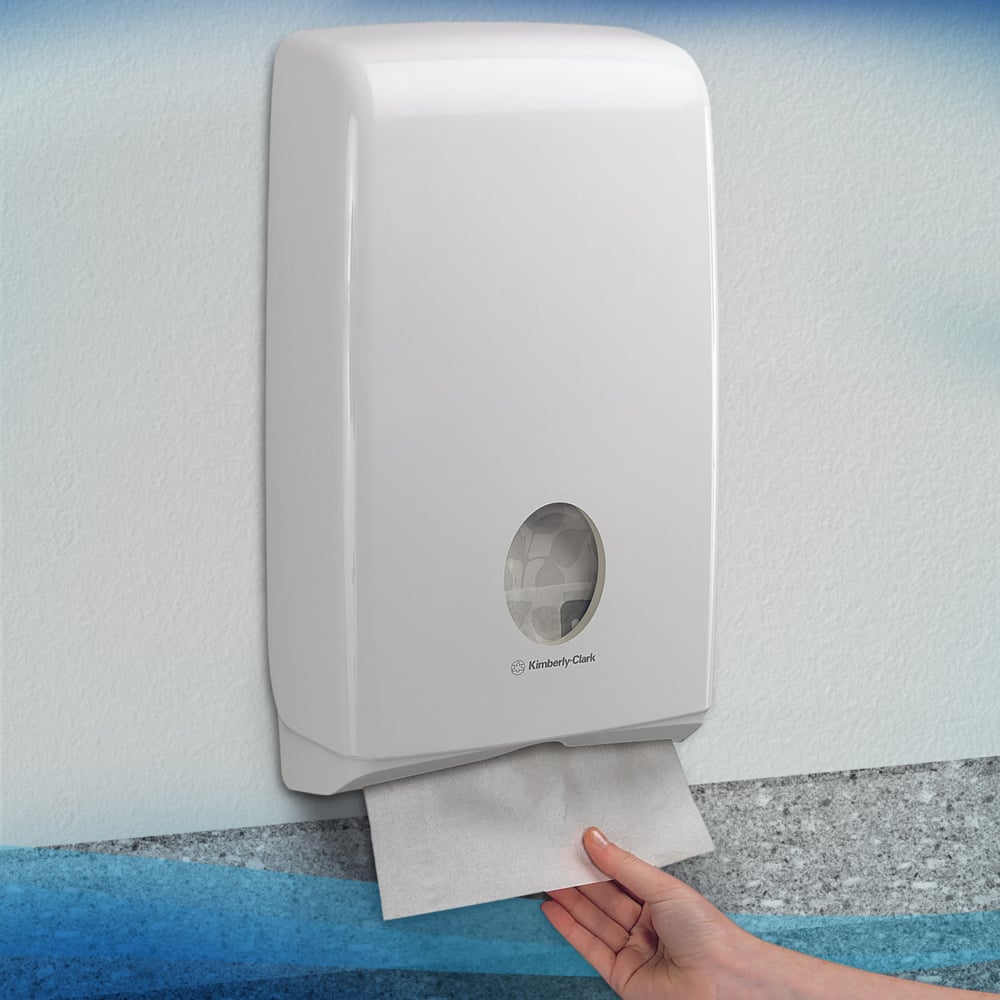 Ręka wyciąga ręcznik Scott® SLIMFOLD z białego dozownika ściennego Kimberly-Clark GmbH z okienkiem, które pokazuje składane ręczniki wewnątrz. Higieniczny dozownik jest zamontowany na jasnej ścianie; mieści 16 paczek x 110 arkuszy na karton.