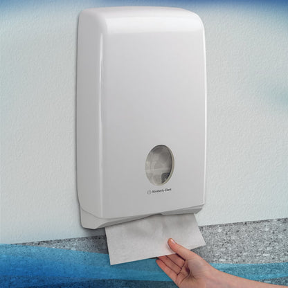 Ręka wyciąga ręcznik Scott® SLIMFOLD z białego dozownika ściennego Kimberly-Clark GmbH z okienkiem, które pokazuje składane ręczniki wewnątrz. Higieniczny dozownik jest zamontowany na jasnej ścianie; mieści 16 paczek x 110 arkuszy na karton.