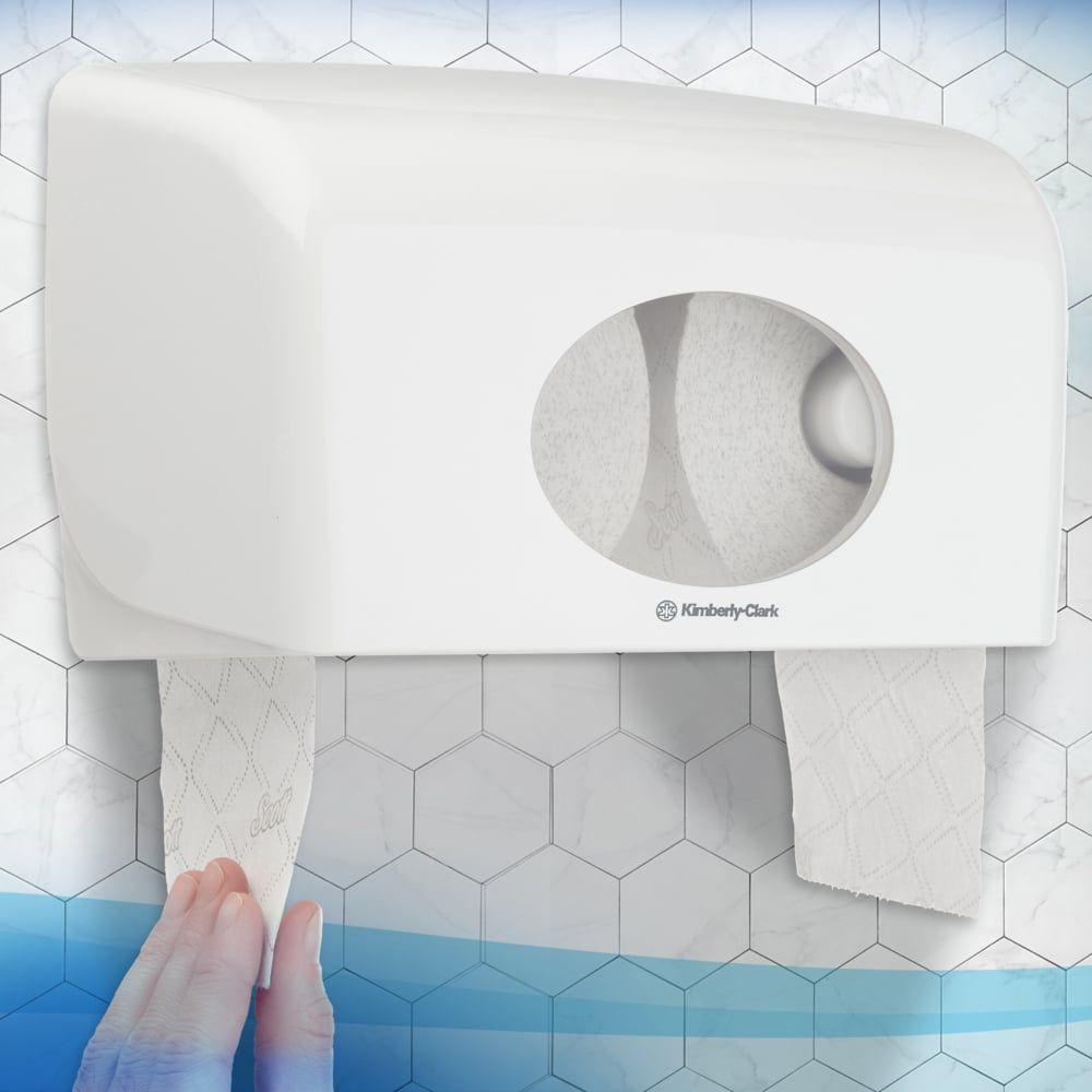 Eine Person zieht FSC-zertifiziertes Scott® Essential™ Toilettenpapier 8517 2-lagig von Kimberly-Clark GmbH aus einem weißen Wandspender vor einer sechseckig gefliesten Badezimmerwand.