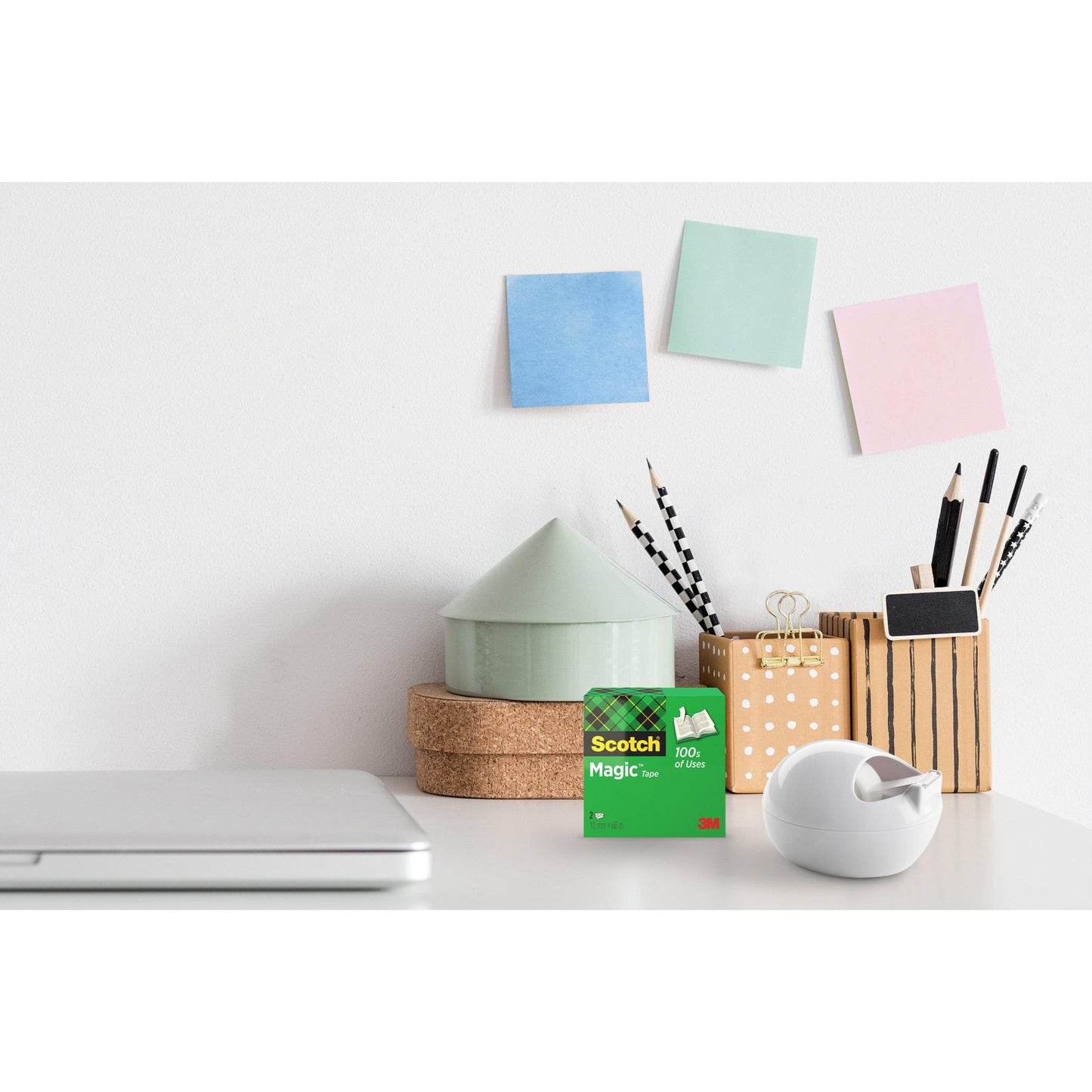 Biurko z zamkniętym laptopem, 3M Deutschland GmbH Scotch™ Karim Rashid Tischabroller i pudełko z taśmą, pojemnik na długopisy i ołówki, mała ozdoba w kształcie domu i trzy kolorowe karteczki samoprzylepne na białej ścianie.