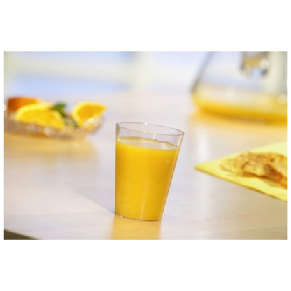 Ein Starpak 50 Trinkbecher, PS glasklar von Starpak, gefüllt mit Orangensaft, steht auf einem hellen Tisch. Dahinter befinden sich Orangenscheiben auf einem Teller und ein verschwommener gelber Gegenstand, möglicherweise Lebensmittel, auf einer gelben Serviette.