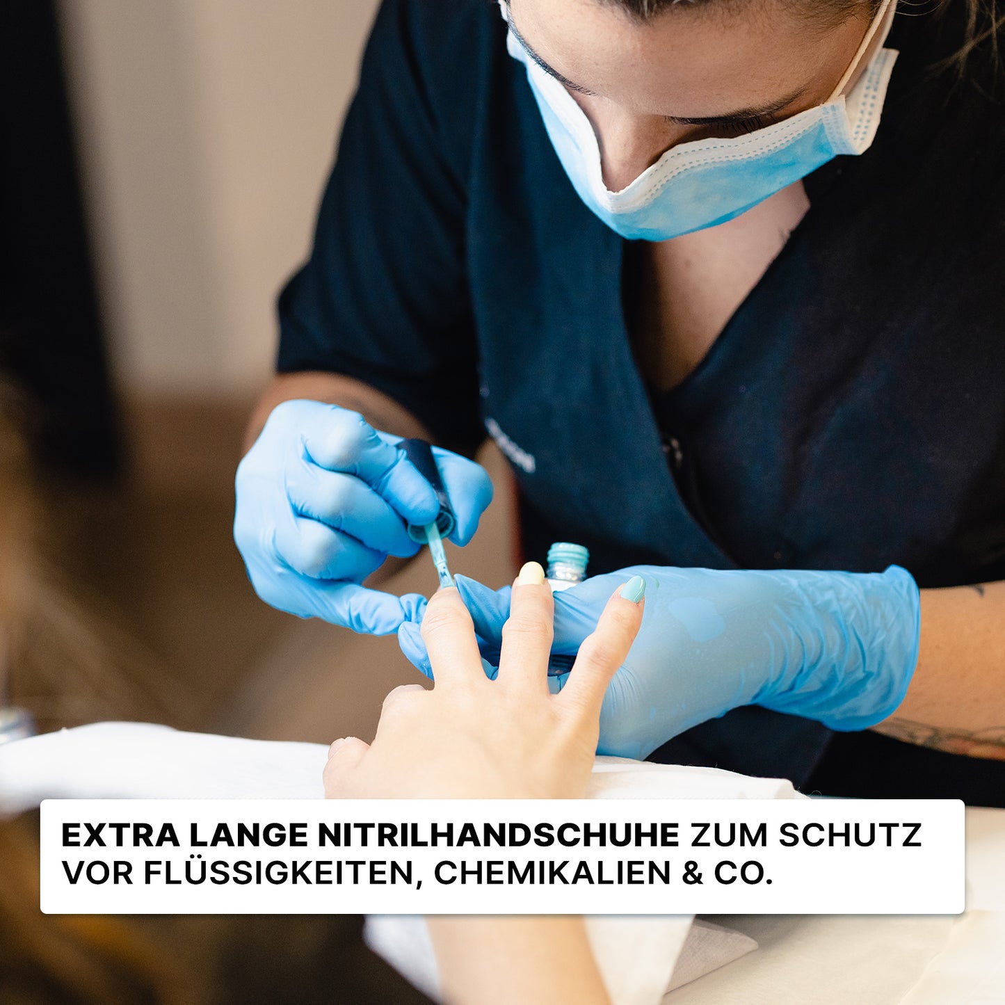 Osoba, która nosi jednorazowe rękawice ARNOMED NITRIL BLUE XTRA LONG (30 cm) firmy ARNOWA GmbH i maskę, maluje komuś paznokcie; tekst niemiecki podkreśla ochronną funkcję niebieskich rękawic nitrylowych.