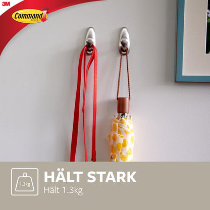 Dwa metalowe haki Command™ "klasyczne" firmy 3M Deutschland GmbH są zamontowane na białej ścianie i trzymają czerwoną smycz oraz żółty wzorzysty parasol. Tekst brzmi: "TRZYMA MOCNO Trzyma 1.3kg" i pokazuje logo Command.