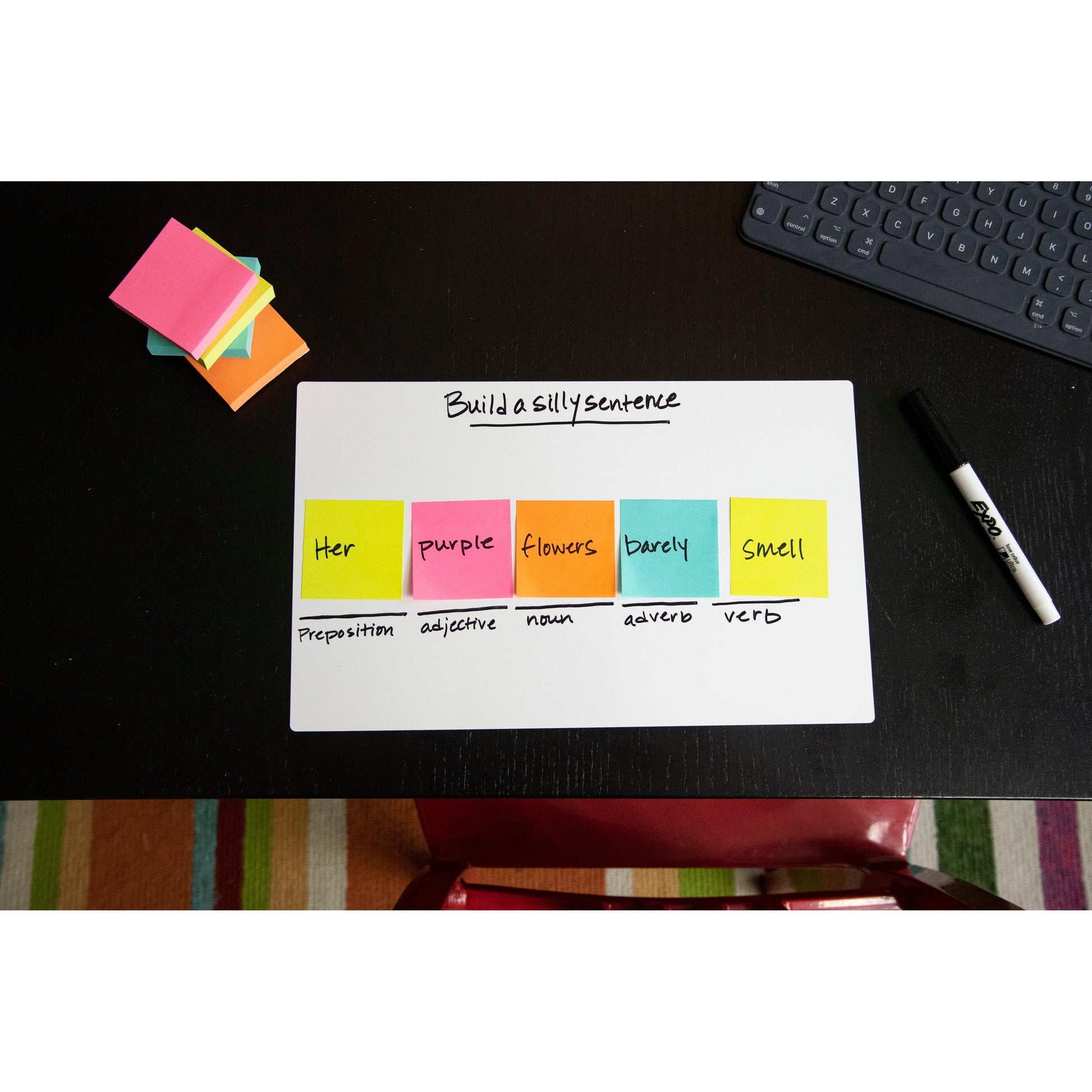 Ein Post-it® Notes Würfel der 3M Deutschland GmbH (76x76mm, 450 Blatt, 100% PEFC) zeigt eine Satzbauaufgabe auf einem Whiteboard mit Grammatiketiketiketten; zusätzliche Haftnotizen, ein Marker, eine Tastatur und ein gestreifter Teppich liegen in der Nähe.