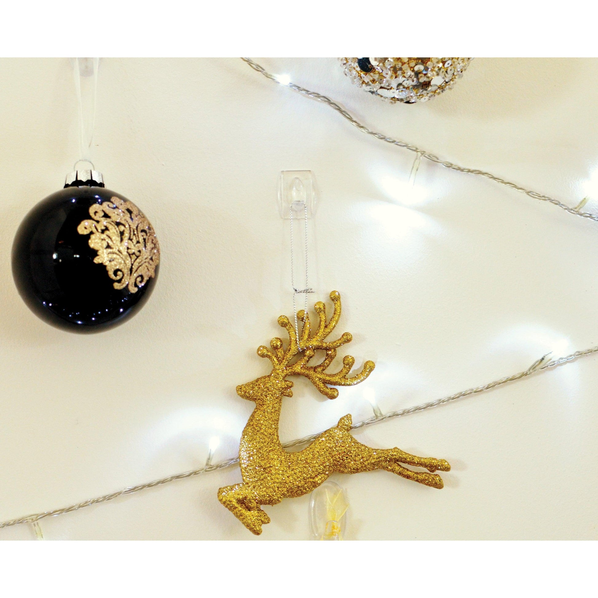 Ein goldenes Glitzer-Rentier, eine schwarze Kugel mit goldenem Dekor und eine gold-weiße Kugel hängen mit dem transparenten Command™ Mini-Haken der 3M Deutschland GmbH an der Wand, während im Hintergrund eine Lichterkette leuchtet.