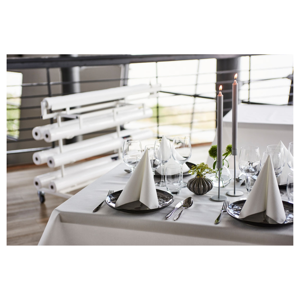Na pięknie nakrytym stole jadalnym stoi świeża, biała rolka obrusu Duni Dunicel® od Duni GmbH, która oferuje materiałopodobną fakturę. Trójkątnie złożone serwetki na ciemnych talerzach towarzyszą kieliszkom do wina. Wysokie świece i małe aranżacje kwiatowe służą jako dekoracja stołu, w tle widać zrolowane płótna.