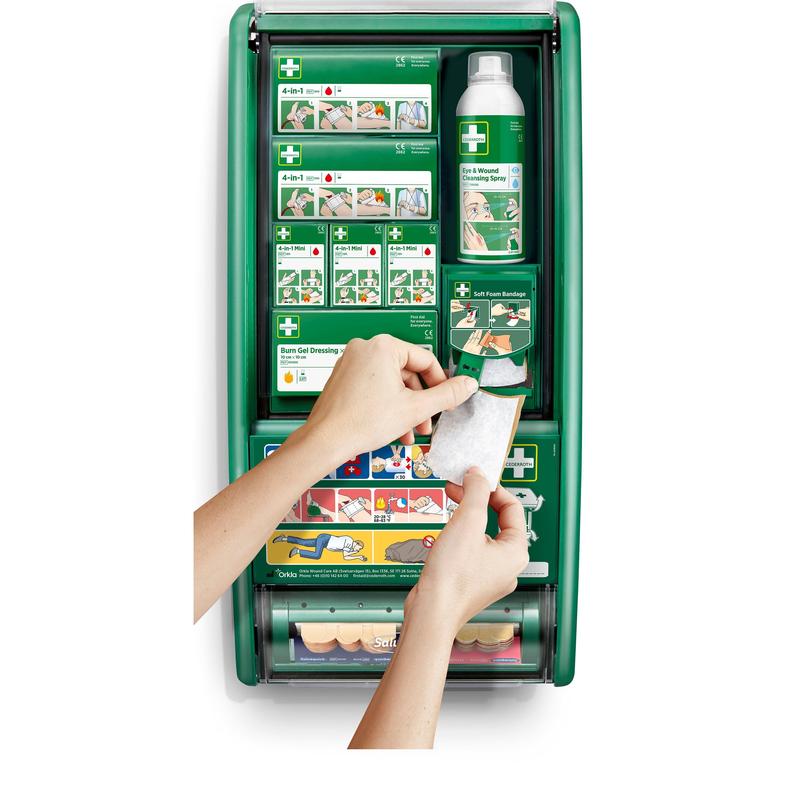 Osoba trzyma bandaż przed stacją pierwszej pomocy Cederroth od Orkla Wound Care AB, która jest wyposażona w bandaże, żel na oparzenia, roztwór soli fizjologicznej i opatrunki. Ilustrowane instrukcje widoczne są na półkach wewnątrz.