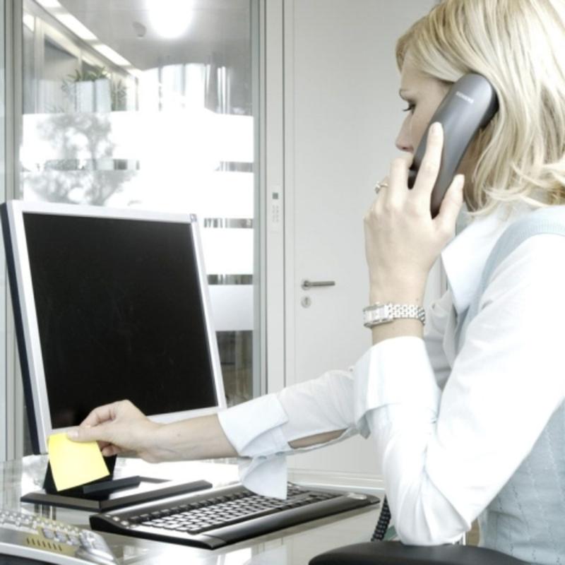 Blond osoba przy biurku kładzie żółtą kostkę notatek samoprzylepnych Post-it® 636-B, 76 mm x 76 mm, od 3M Deutschland GmbH (450 arkuszy, PEFC) na monitorze podczas rozmowy telefonicznej. Na stole leży również klawiatura, mysz i komputer.