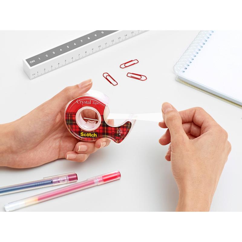 Osoba wyciąga taśmę klejącą Scotch® Crystal (19 mm x 7,5 m) z czerwonego podajnika firmy 3M Deutschland GmbH na białym biurku z czerwonymi spinaczami, długopisami, linijką i spiralnym notatnikiem.