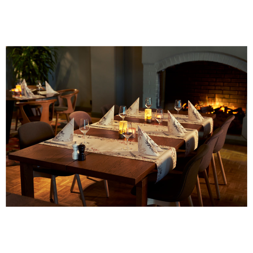 Ein gemütlicher Essbereich verfügt über zwei Holztische mit Weingläsern und Duni Bio Dunisoft® Servietten neben flackernden Kerzen. Dunkle Stühle umgeben die Tische, aufgewertet durch einen brennenden Kamin. Plüschige Sitzgelegenheiten und eine Pflanze tragen zum naturinspirierten Design bei.