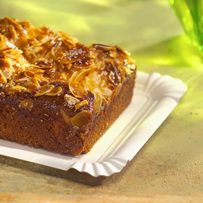 Na prostokątnym talerzu papierowym PAPSTAR, 13x20 cm, biały, "pure" na uroczystości (12 sztuk) firmy PAPSTAR GmbH spoczywa prostokątne ciasto z płatkami migdałów - ekologiczna, elegancka opcja serwowania na jasnej powierzchni z zielonym szkłem w tle.