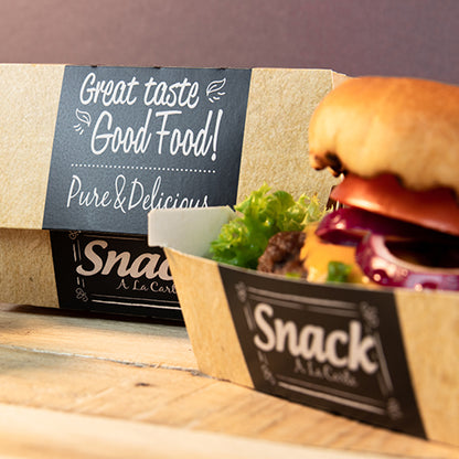 Tacka z tektury z napisem "Snack A La Carte" zawiera hamburgera z sałatą, pomidorem i cebulą. W tle znajduje się PAPSTAR 50 pudełek na burgery, tektura "pure" "Good Food" od PAPSTAR GmbH: Dobry smak Good Food! Czyste i pyszne.