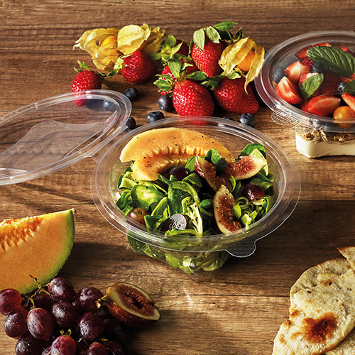 Eine Starpak 50 Feinkost- und Salatschale mit Klappdeckel, R-PET glasklar rund, hält einen Melonen- und Feigensalat auf einem Holztisch mit Erdbeeren, Weintrauben, Brot, Kirschen und Obst. Ein weiterer Starpak R-PET-Behälter mit Obst steht in der Nähe.