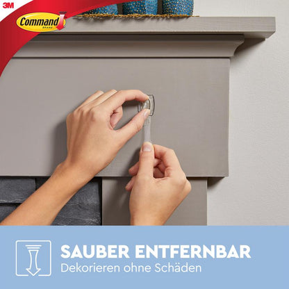 Osoba zdejmuje przezroczysty pasek klejący z szarego gzymsu kominkowego i pokazuje, jak przezroczyste mini-haczyki Command™ z przezroczystymi paskami (17006CLR) od 3M Deutschland GmbH dekorują i mocują bez śladów. Widoczne są loga Command i 3M.