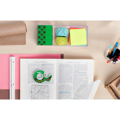 Biurko z otwartą książką, zestaw promocyjny Scotch® Magic™ Invisible Tape (24 rolki, 3M Deutschland GmbH), kolorowy papier, karteczki samoprzylepne, sznurek, gumka, kubek z pisakami i ołówkami oraz stosy książek i zwinięty brązowy papier.