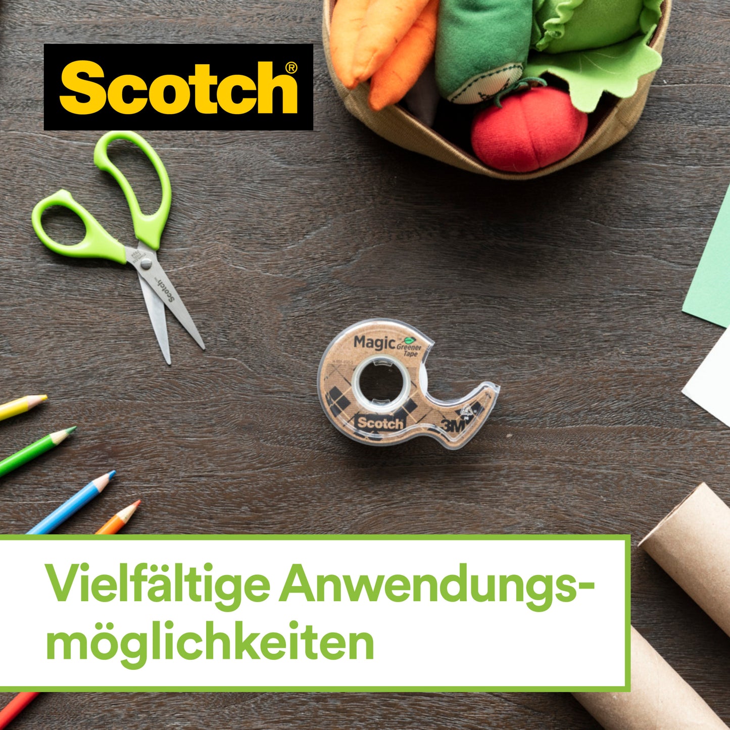 Eine Schere mit grünem Griff, Buntstifte, grünes Papier, Pappröhren, Stoffvegetation in einem Korb und der Scotch™ Edelstahl-Klebebandabroller mit Scotch® Magic™ Klebeband (66% pflanzenbasiert) von 3M auf einem Holztisch. Vielfältige Anwendungsmöglichkeiten.