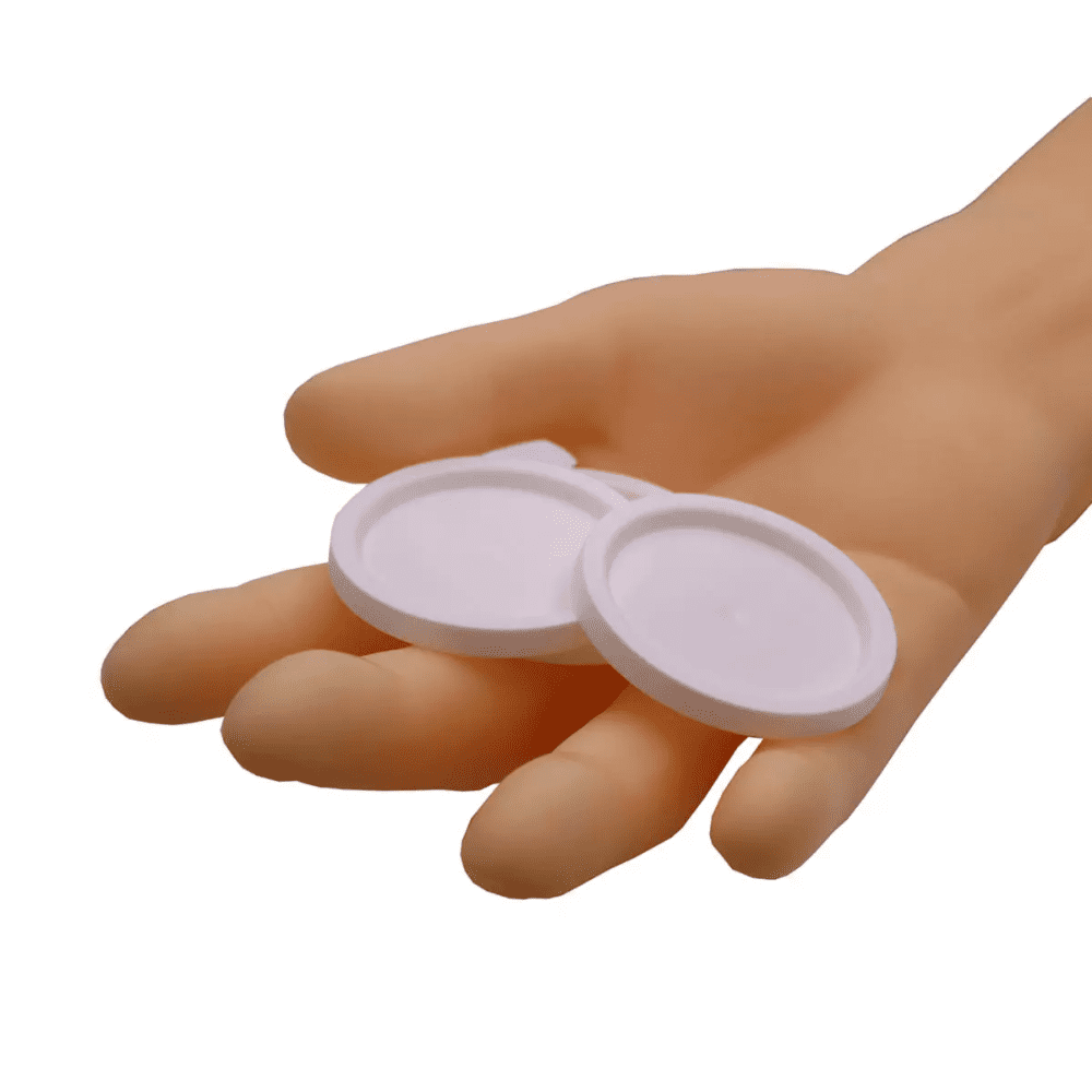 Ręka trzyma dwa okrągłe pokrywki AMPri do kubków medycznych, białe obiekty na prostym białym tle.