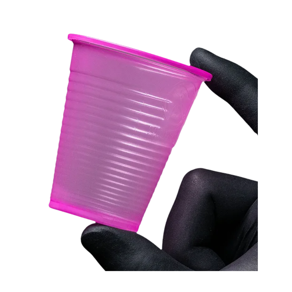 Mały, różowy kubek do płukania ust AMPri Dental PP z polipropylenu jest trzymany między dwoma rękawiczkami na tle białym. Ten jednorazowy kubek do płukania ust AMPri Handelsgesellschaft mbH, często spotykany w gabinetach stomatologicznych, ma teksturowaną powierzchnię z poziomymi rowkami.