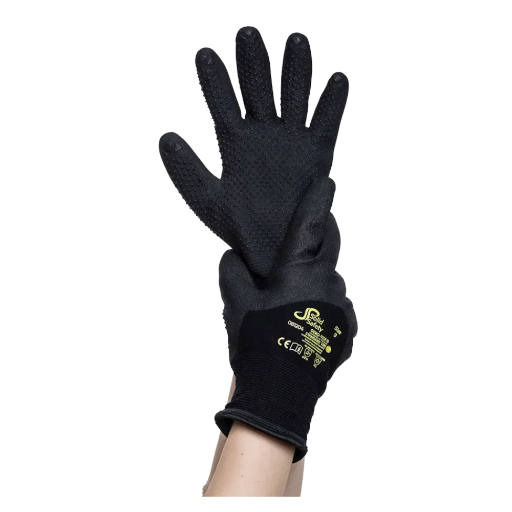 Para czarnych rękawic ochronnych AMPri SolidSafety Tough Winter Dots odpornych na zimno z teksturowanymi dłońmi jest trzymana w górze przez rękę. Te rękawice, które mają poprawiać zdolności manualne, noszą na odwrocie żółty znak towarowy i znaki certyfikacji bezpieczeństwa AMPri Handelsgesellschaft mbH.