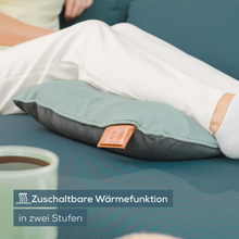 Osoba siedzi na kanapie, jej nogi są wyciągnięte i spoczywają na niebieskoszarej Beurer poduszce masującej MG 139 od Beurer GmbH. Poduszka ma małą jasnobrązową etykietę i naklejkę z napisem „Zuschaltbare Wärmefunktion in zwei Stufen”, co wskazuje, że poduszka posiada regulowane funkcje grzewcze.