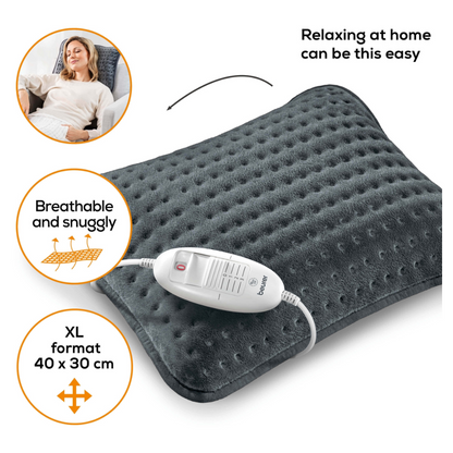 Kobieta leży na sofie i uśmiecha się z zamkniętymi oczami. Obok niej znajduje się Beurer HK 48 poduszka grzewcza na sofę Cosy w ciemnoszarym kolorze z białą jednostką sterującą. Na poduszce widnieje napis „Tak łatwo można się zrelaksować w domu”, „Oddychająca i przytulna” oraz „Format XL 40 x 30 cm”.