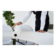 Osoba w wieczorowym stroju, z białą koszulą i czarną kamizelką, rozwija rolkę obrusów Dunicel (1,18 m x 40 m) od Duni GmbH na stole; na prostym tle widać trochę zieleni.