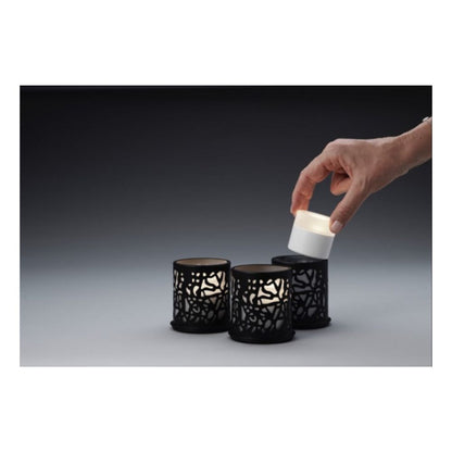 Eine Hand hebt eine kleine weiße LED-Kerze aus einer schwarzen Halterung, während zwei identische Duni GmbH Multicolour LED Set, Mini - 8 Stück in ähnlichen Halterungen auf einer grauen Fläche vor dunklem Hintergrund ruhen.