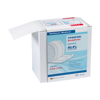 Przedstawione jest pudełko plastra opatrunkowego Holthaus Ypsipor z włókniny firmy Holthaus Medical GmbH & Co. KG. Na pudełku jest napisane, że jest delikatne, ma wymiary 6 cm x 5 m i jest produkowane w Niemczech. Część rolki przyjaznego dla skóry plastra z włókniny jest widoczna i wystaje z pudełka.