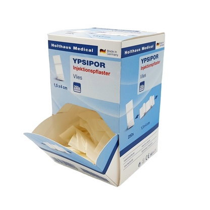 Prostokątne pudełko plastrów iniekcyjnych Holthaus Ypsipor od Holthaus Medical GmbH & Co. KG z napisem „Made in Germany“. Pudełko jest przeważnie jasnoniebieskie i białe, ma wymiary 1,5 x 4 cm, zawiera 250 plastrów z włókniny z wkładką chłonną i jest przedstawione z otwartą górną klapką, która ukazuje zawartość.