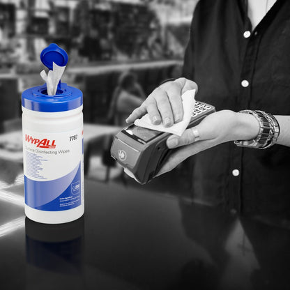 Ktoś używa chusteczki do dezynfekcji powierzchni WypAll® (Kimberly-Clark GmbH) z otwartej wanny dozującej do dezynfekcji terminala kart płatniczych. Biały pojemnik (200 chusteczek) stoi na ladzie, tło jest rozmyte.